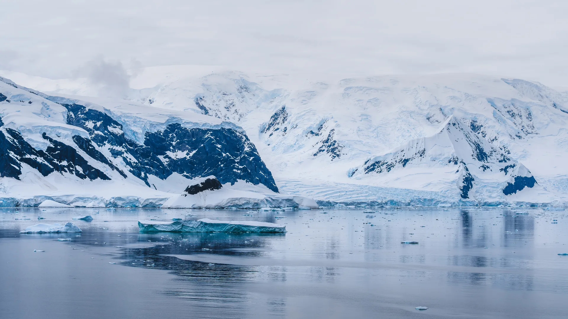 Un témpano bajo flotando en agua quieta frente a un frente de glaciar y montañas nevadas